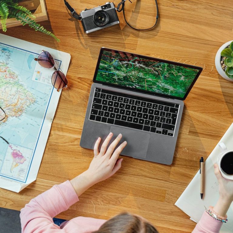 A person using a laptop on a wooden table with a map, camera, and sunglasses nearby.
