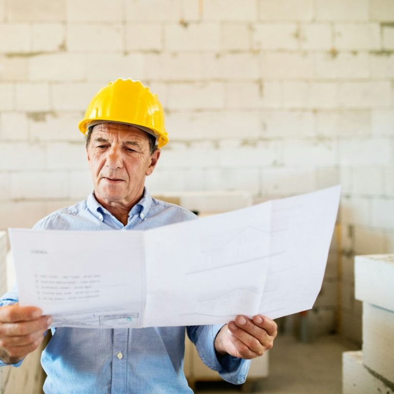 An older man in a yellow hard hat reviews blueprints at a construction site.
