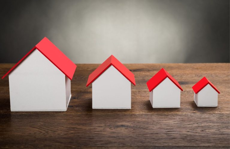 Downsizing illustration image Four model houses of varying sizes, all with white walls and red roofs, on a wooden surface.