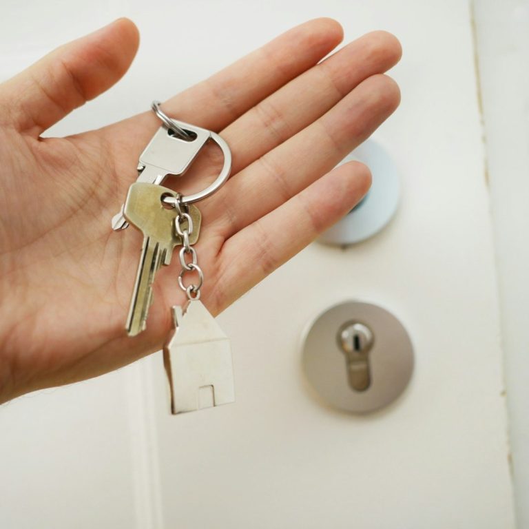 Hand holding keys with a house keychain, positioned near a door lock. 
