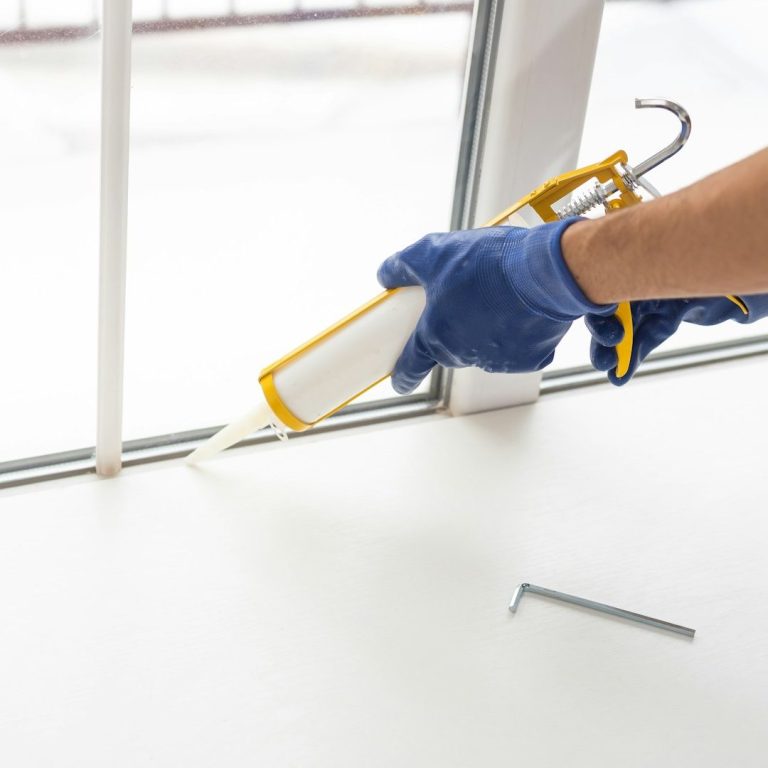 A man freshening up a home for sale A hand applying sealant with a caulking gun at a window frame.